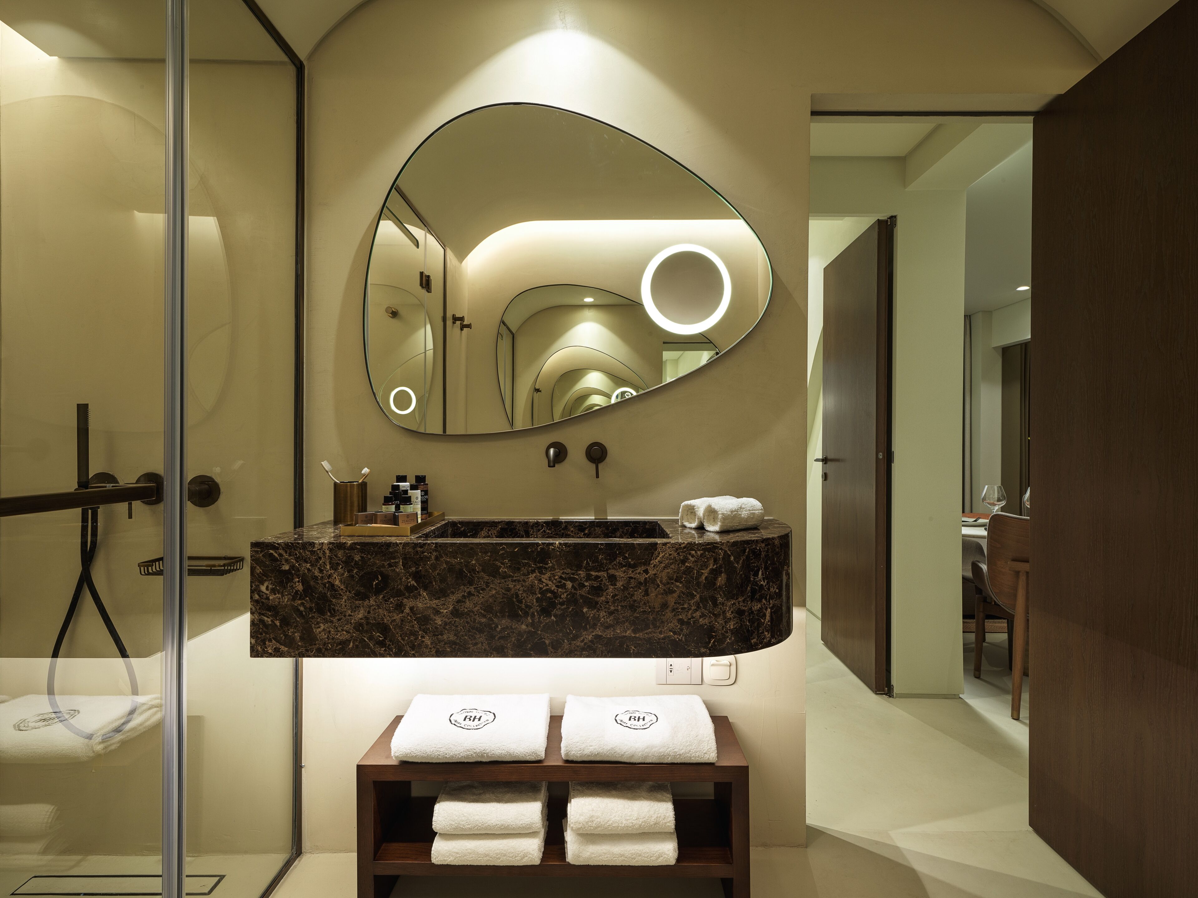 Une salle de bain moderne avec un lavabo en marbre, un grand miroir doté d’un éclairage circulaire et des serviettes soigneusement pliées, rangées sur une étagère.