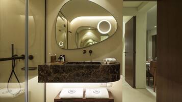 Une salle de bain moderne avec un lavabo en marbre, un grand miroir doté d’un éclairage circulaire et des serviettes soigneusement pliées, rangées sur une étagère.