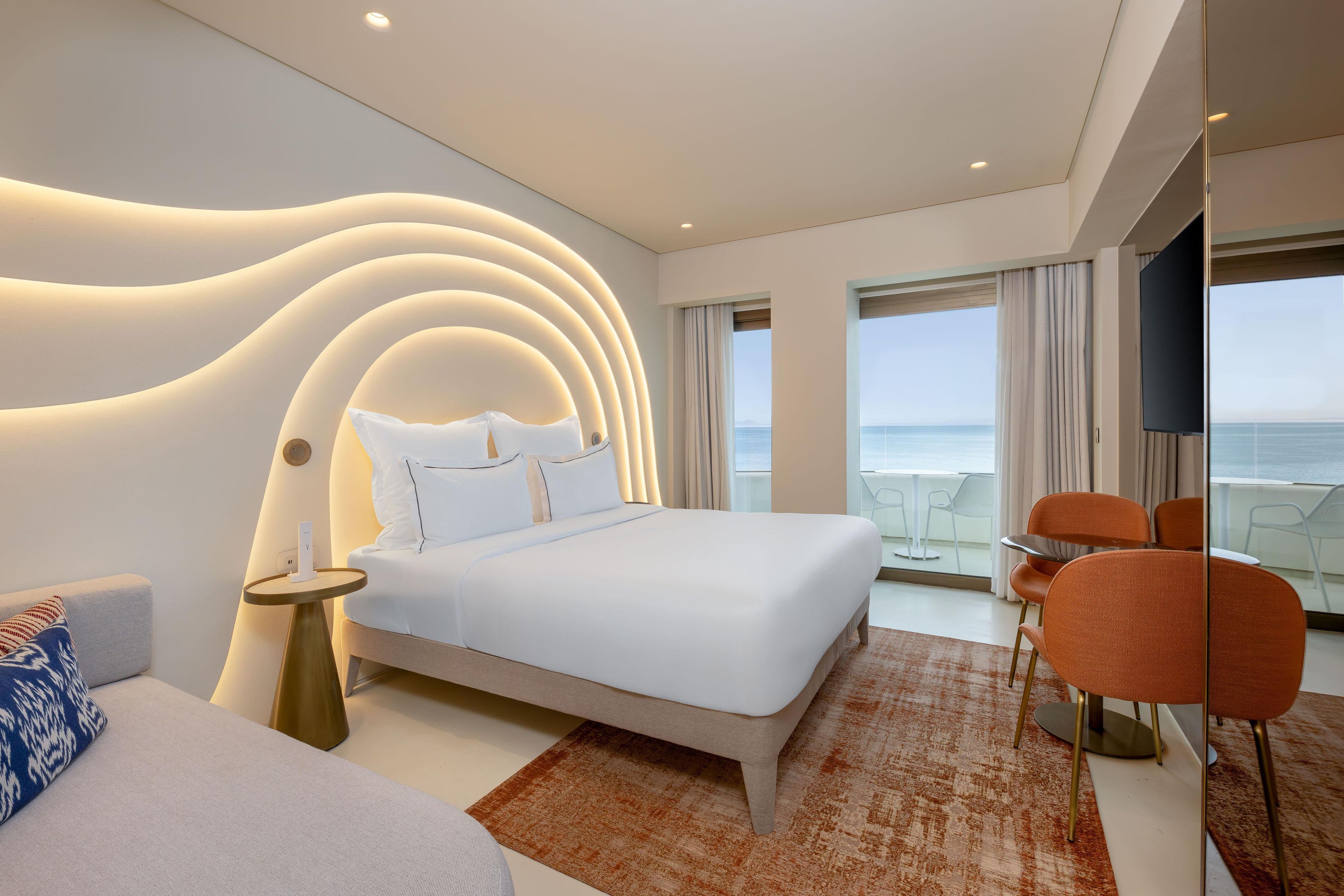 A modern hotel room with a large bed, a seating area with a view of the sea, and a balcony with outdoor furniture.