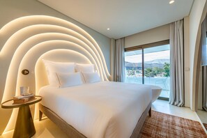A modern hotel room with a large bed, a round side table, and a view of the city through the window. - Isla Brown Corinthia Resort & Spa, Curio Collection Hilton (Loutraki-Agioi Theodoroi)