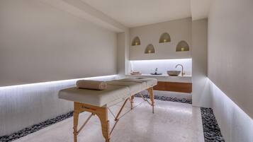 A spa room with a massage table, a small sink, and a window with three lights.
