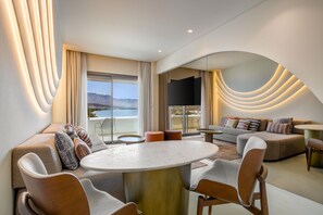 A modern living room with a round table, sofas, and a large window offering a view of the mountains. - Isla Brown Corinthia Resort & Spa, Curio Collection Hilton (Loutraki-Agioi Theodoroi)
