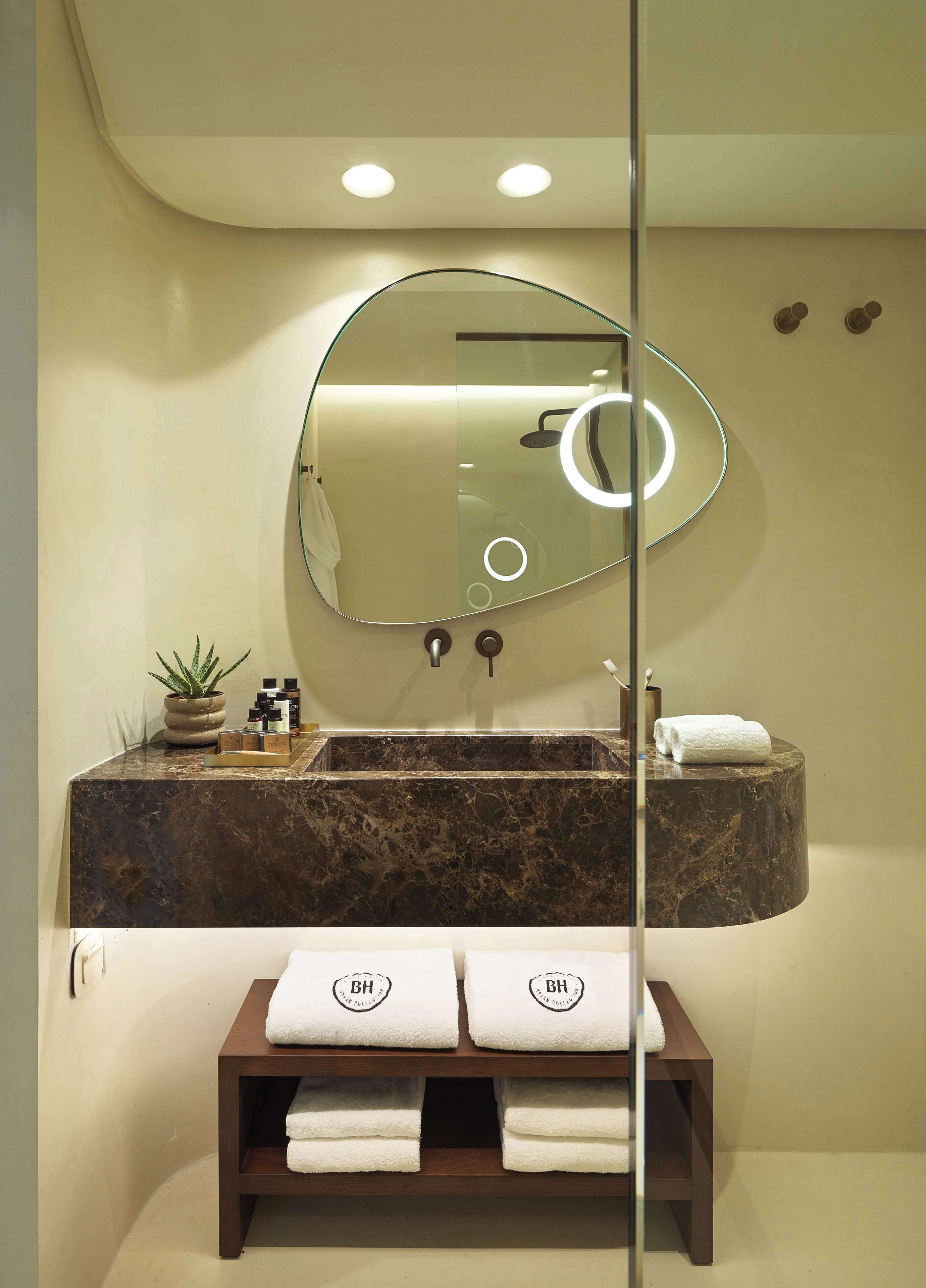 A bathroom with a marble sink, a large mirror with a circular light, a small potted plant, and a wooden shelf with towels.