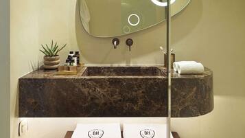 A bathroom with a marble sink, a large mirror with a circular light, a small potted plant, and a wooden shelf with towels.