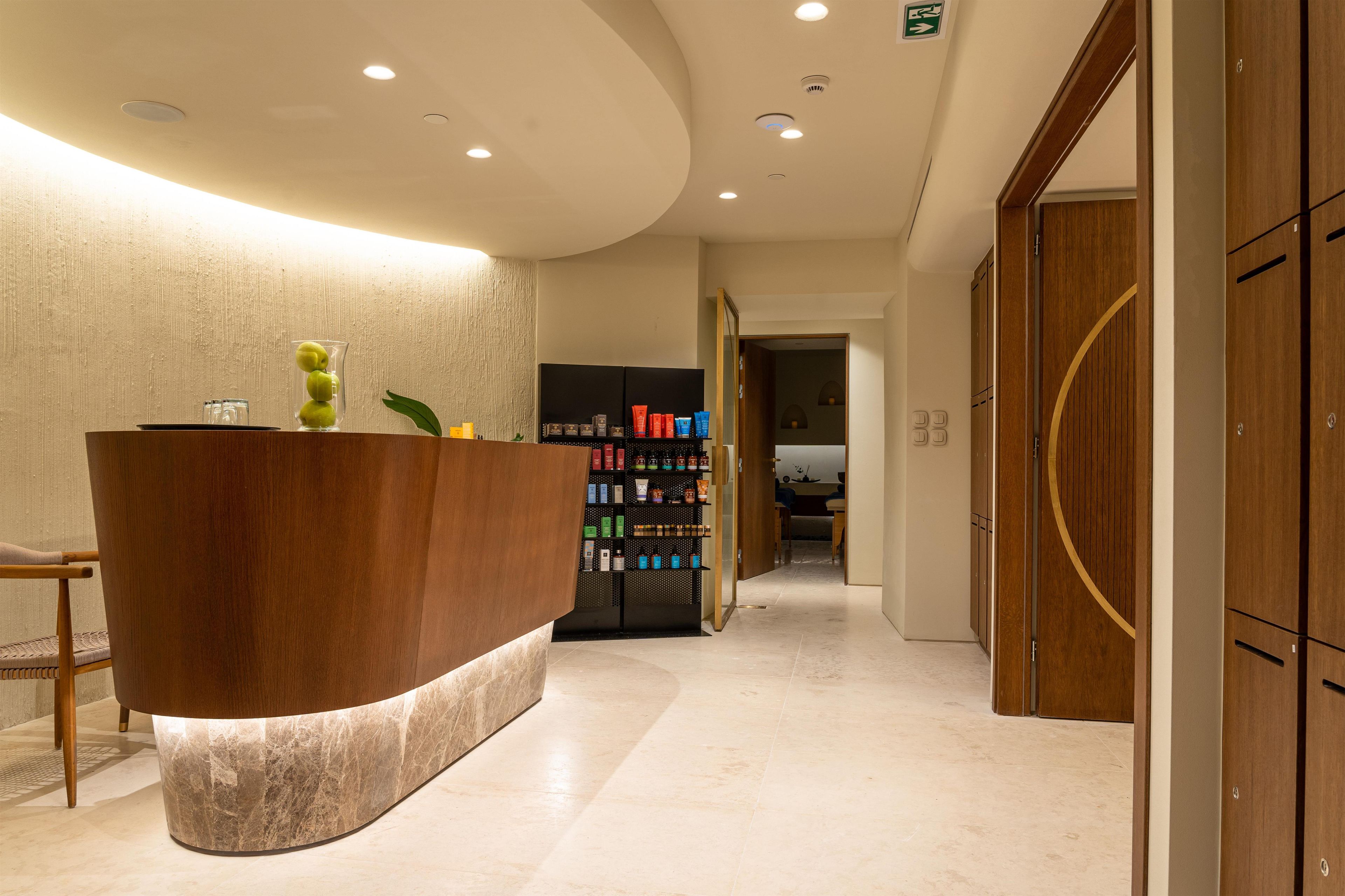 A hotel reception area with a curved wooden reception desk, a wall-mounted shelf with toiletries, and a hallway leading to a room.