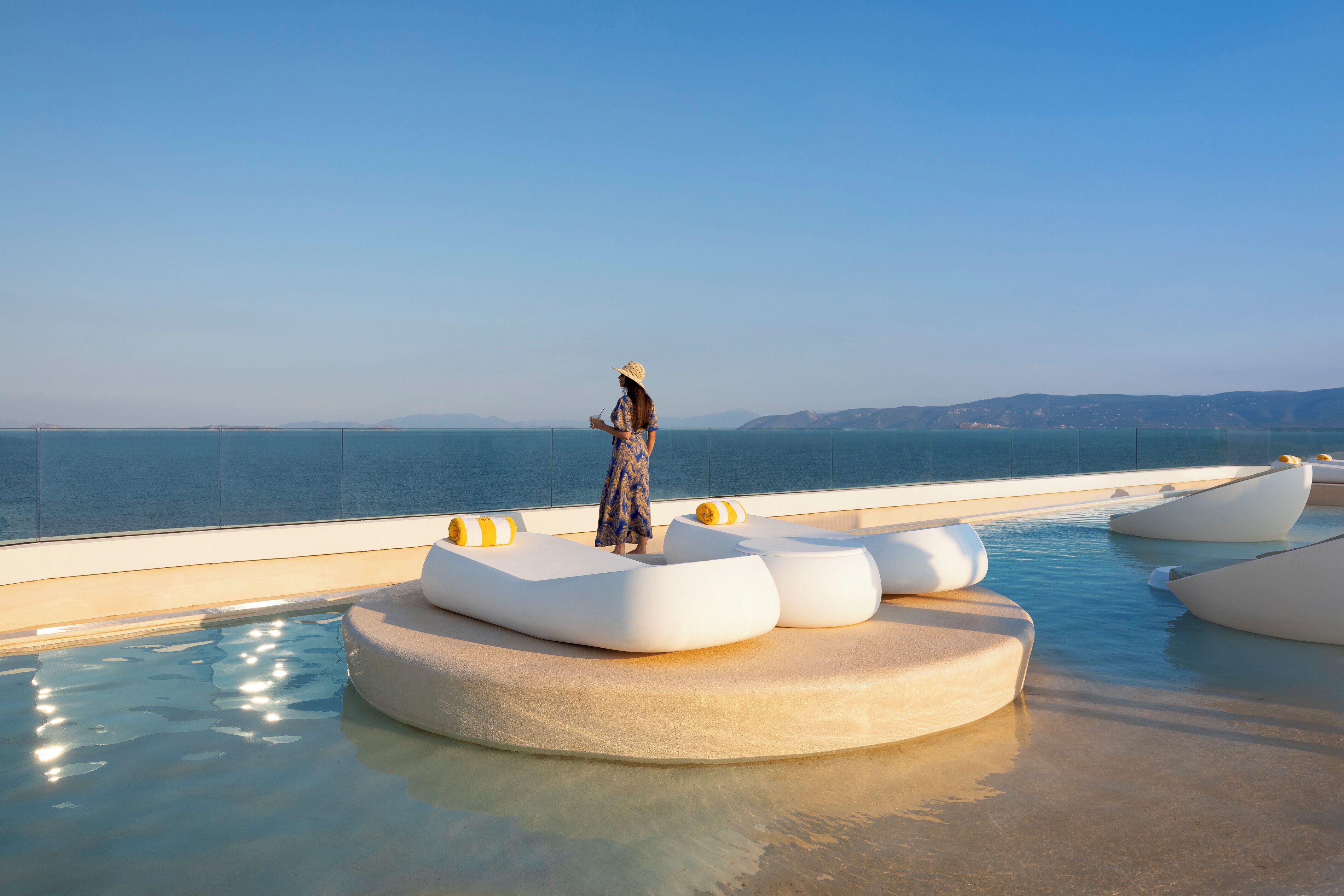 Una persona está de pie sobre un moderno borde de piscina con asientos blancos y amarillos originales, con vistas al mar azul cristalino y colinas lejanas.