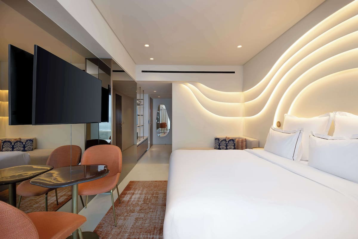 Une chambre d’hôtel moderne dotée d’un grand lit, d’un téléviseur fixé au mur, d’un coin repas avec des chaises et d’une salle de bain visible à travers une porte en verre.
