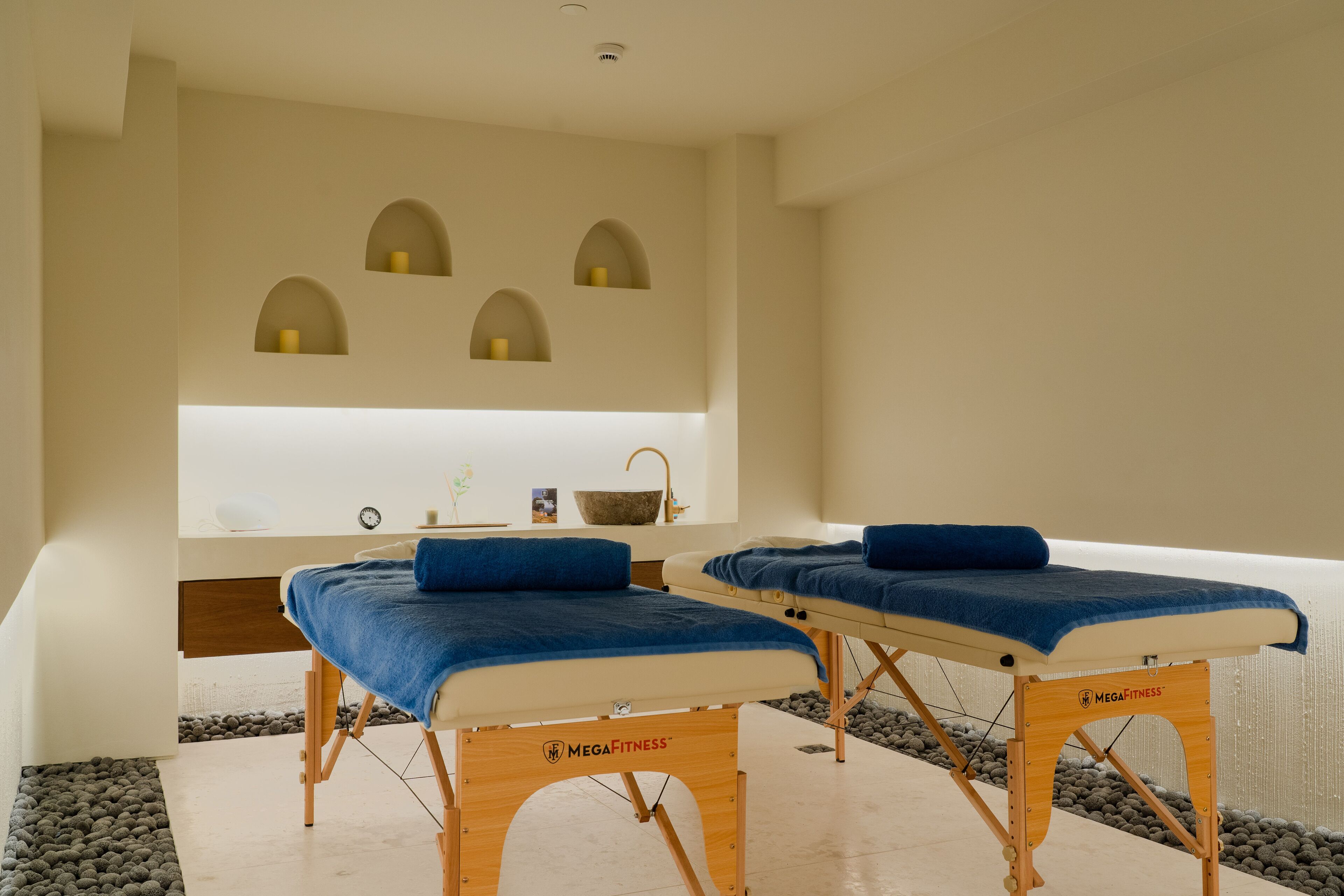 Une salle de spa équipée de deux tables de massage, d’un plancher en bois, d’un mur en pierre et d’une étagère où sont posées des bougies et un bol.