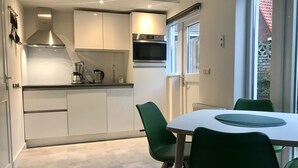 Een moderne keuken met witte kasten, een roestvrijstalen afzuigkap en een inbouwoven. Er is een eettafel met groene stoelen en een raam met uitzicht op een dak met rode dakpannen.