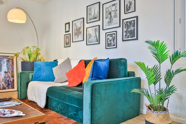 Una sala de estar con un sofá verde, cojines coloridos, una planta grande y cuadros en la pared.