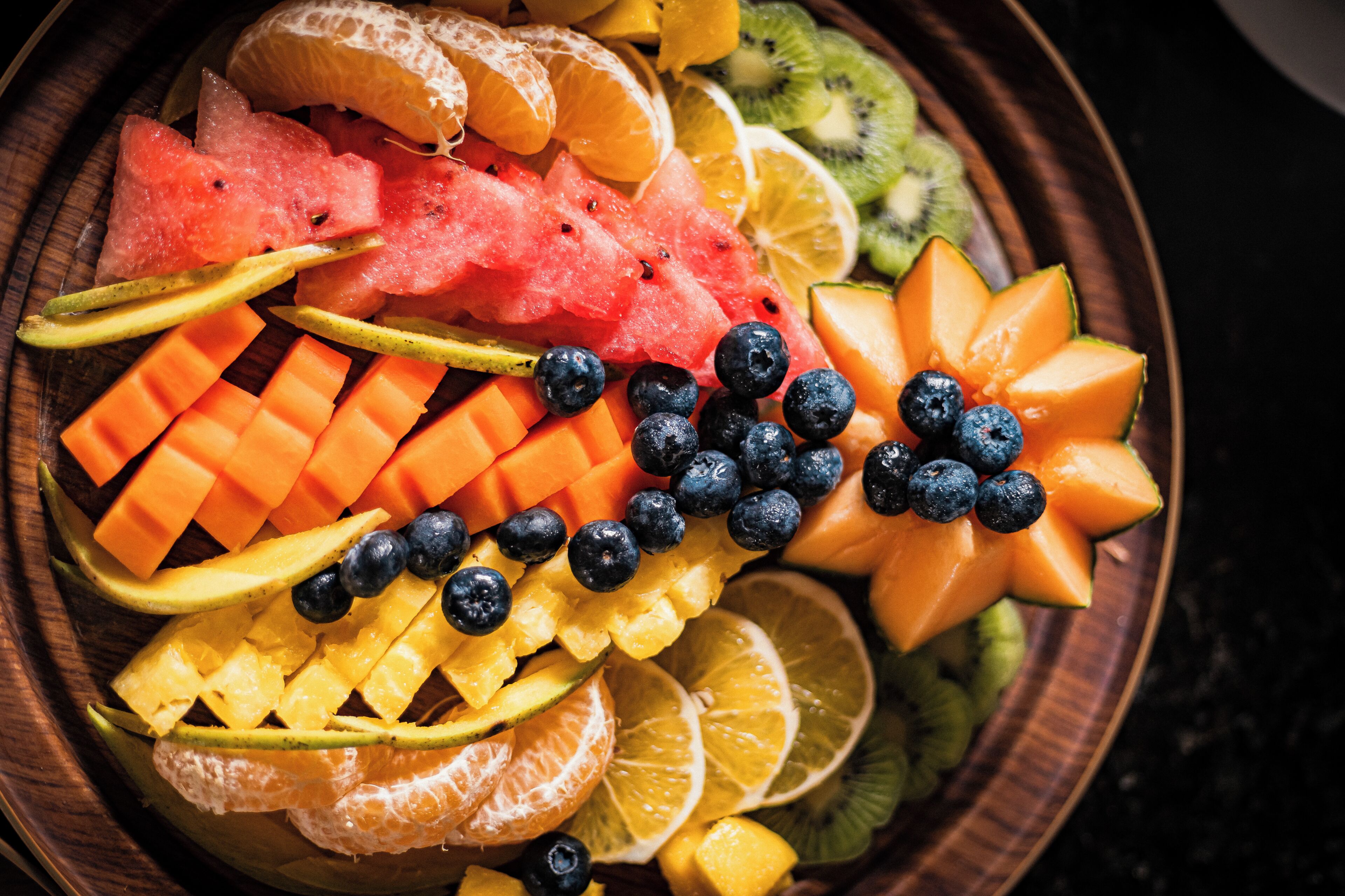 Un saladier en bois rempli de divers fruits, notamment de la pastèque, des oranges, des kiwis, des myrtilles et des melons.