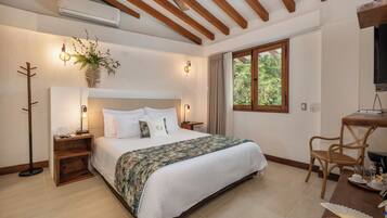 A bedroom with a large bed, wooden ceiling beams, a bedside table with a lamp, a potted plant, and a window with curtains.