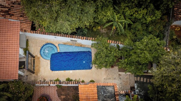 An aerial view of a rooftop pool area with a large rectangular pool, a smaller circular pool, and surrounding greenery.