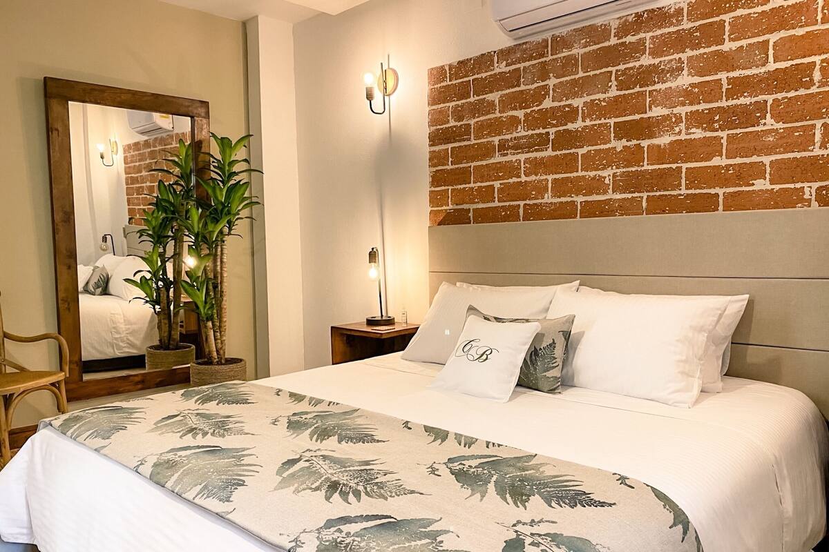 Una camera d'albergo con un letto, uno specchio, un comodino, una lampada e un muro in mattoni.