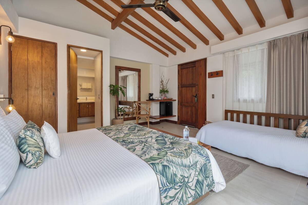 Una camera d'albergo con due letti, travi a vista sul soffitto, un bagno visibile attraverso una porta aperta e una scrivania con una sedia.