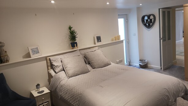 Une chambre avec un grand lit, une table de chevet, une chaise, une plante en pot et un miroir en forme de cœur accroché au mur.
