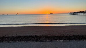 Une plage avec une jetée qui s’avance dans l’eau, un coucher de soleil en arrière-plan et une mer calme.