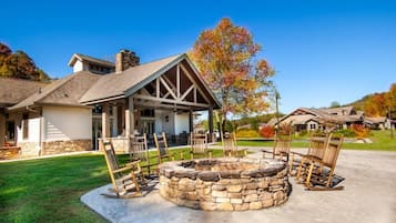 Un foyer en pierre, entouré de fauteuils à bascule en bois, avec une terrasse couverte et une maison en arrière-plan.