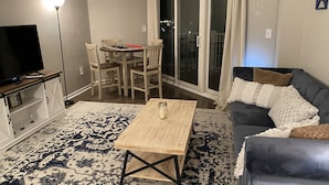 En stue med fjernsyn, sofabord, sofa og et spiseområde med stole og bord.