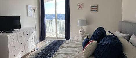 A bedroom with a bed, a TV on a dresser, a nightstand with a lamp, and a view of the outside through a window.