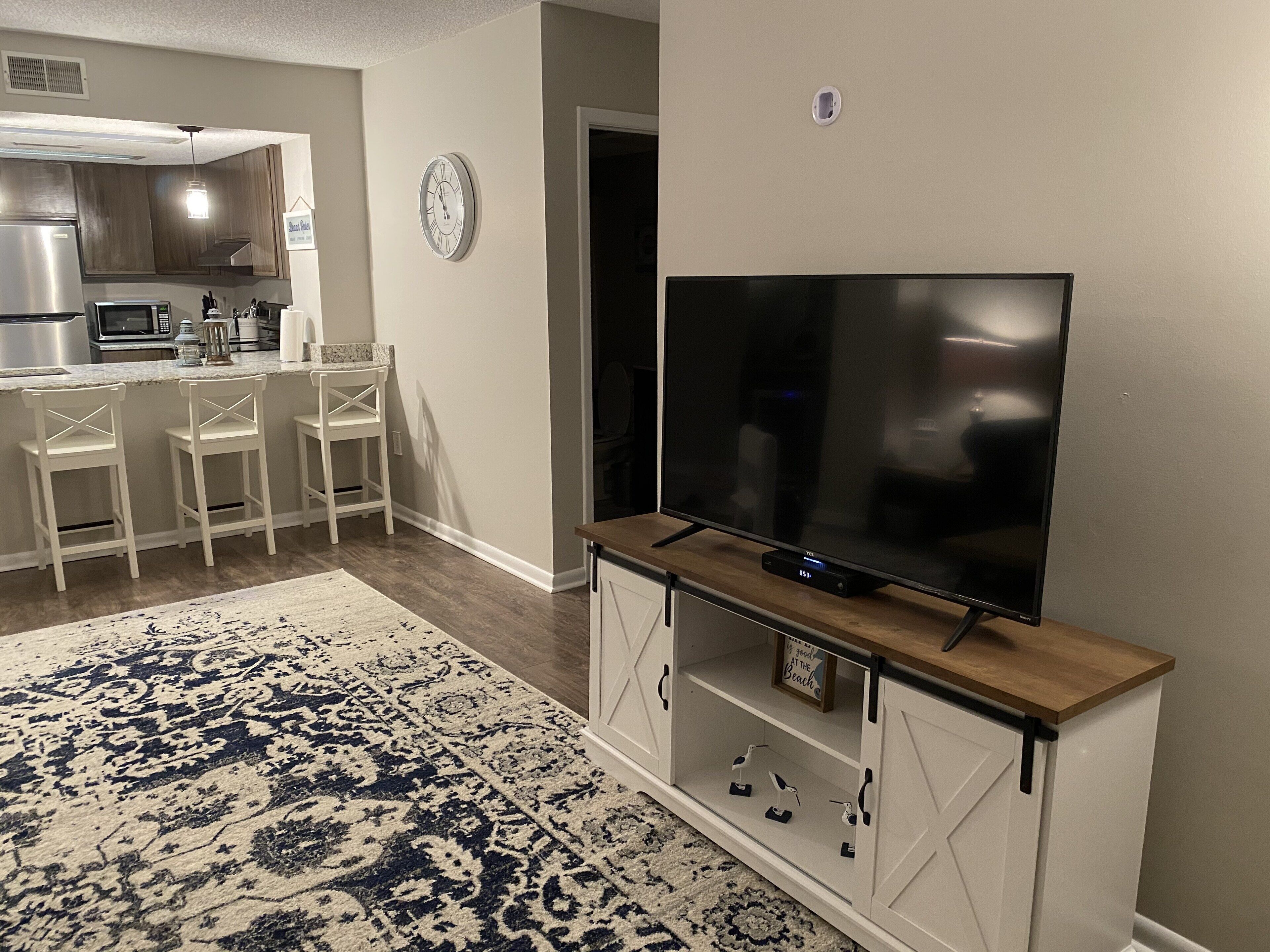 Una sala de estar moderna con una televisión de pantalla plana sobre un soporte de madera, una alfombra estampada y una cocina al fondo.