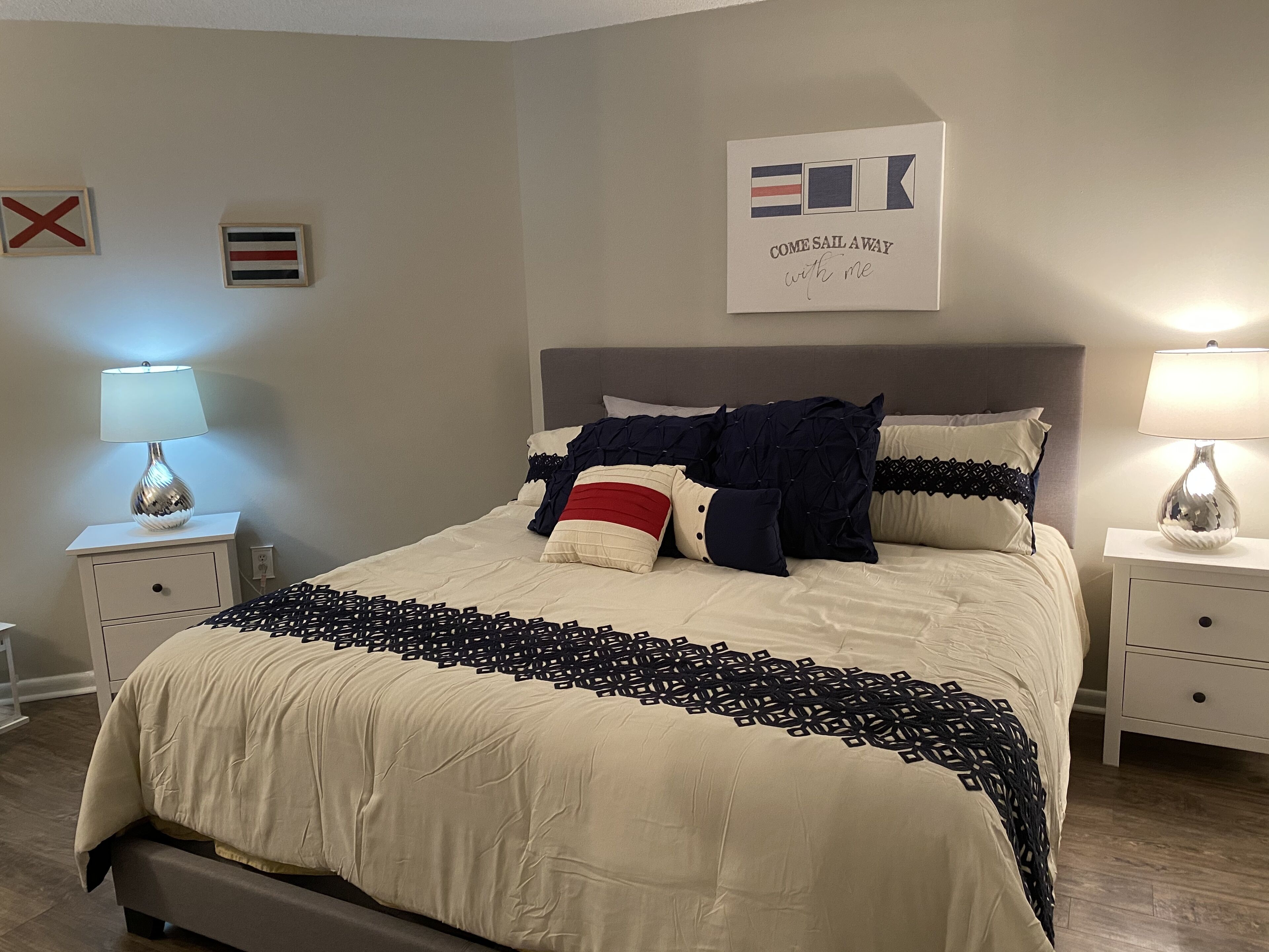Una cama bien hecha con una colcha estampada, dos lámparas de noche y una pared con una bandera y un cartel que dice 'COME SAIL AWAY with me'.