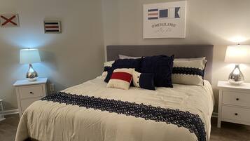 Una cama bien hecha con una colcha estampada, dos lámparas de noche y una pared con una bandera y un cartel que dice 'COME SAIL AWAY with me'.