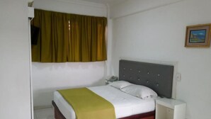 Una cama individual con cabecera gris y colchón blanco, una manta verde y una mesita de noche con un teléfono.