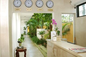 Un área de recepción con relojes que muestran la hora de Nueva York, Londres y Tokio, un mostrador de recepción y un exuberante jardín interior.