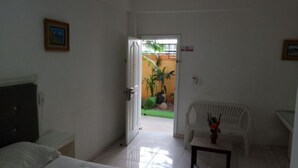 Habitación de hotel con cama, escritorio, silla y una mesita. Se ve una puerta que da a un patio con plantas y una valla.