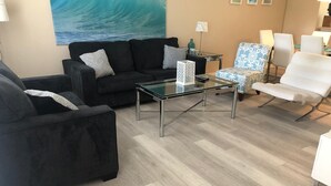Uma sala de estar moderna com uma mesa de centro de vidro, sofás escuros e poltronas estampadas.