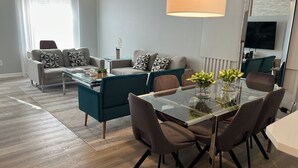 Uma sala de estar moderna com uma mesa de jantar de vidro, cadeiras verde-água e marrom, um sofá cinza com almofadas estampadas e uma televisão de tela plana fixada na parede.