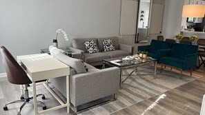 A modern living room with a grey sofa, a glass coffee table, a white desk, and teal armchairs.