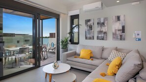 A modern living room with a grey sofa, yellow cushions, a small white table, and a glass door leading to an outdoor dining area. - Porti Iraklis 4 - A Stylish Eco Villa near beaches (Rethymno)