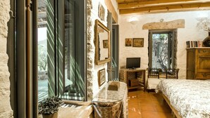 A rustic bedroom with a wooden ceiling, a bed with a patterned bedspread, a small table with a vase, and a window with a view of trees. - Dolly's Dream House (Patmos)
