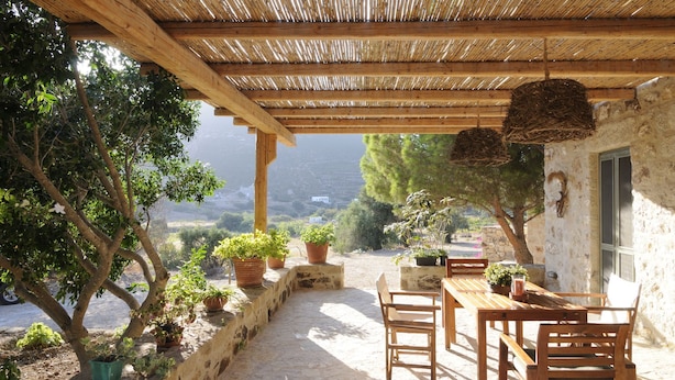 A covered outdoor patio with wooden furniture and potted plants, overlooking a scenic landscape. - Dolly's Dream House (Patmos)