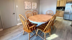 A dining area with a round wooden table and six chairs, a kitchen with wooden cabinets and a stainless steel refrigerator, and two wall-mounted artworks. - Little Bass Cabin - Kayaks & Clear Water Swimming (Grand Rapids)