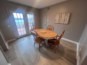 A dining area with a wooden table and chairs, a window with blinds, and a wall-mounted artwork. - Little Bass Cabin - Kayaks & Clear Water Swimming (Grand Rapids)