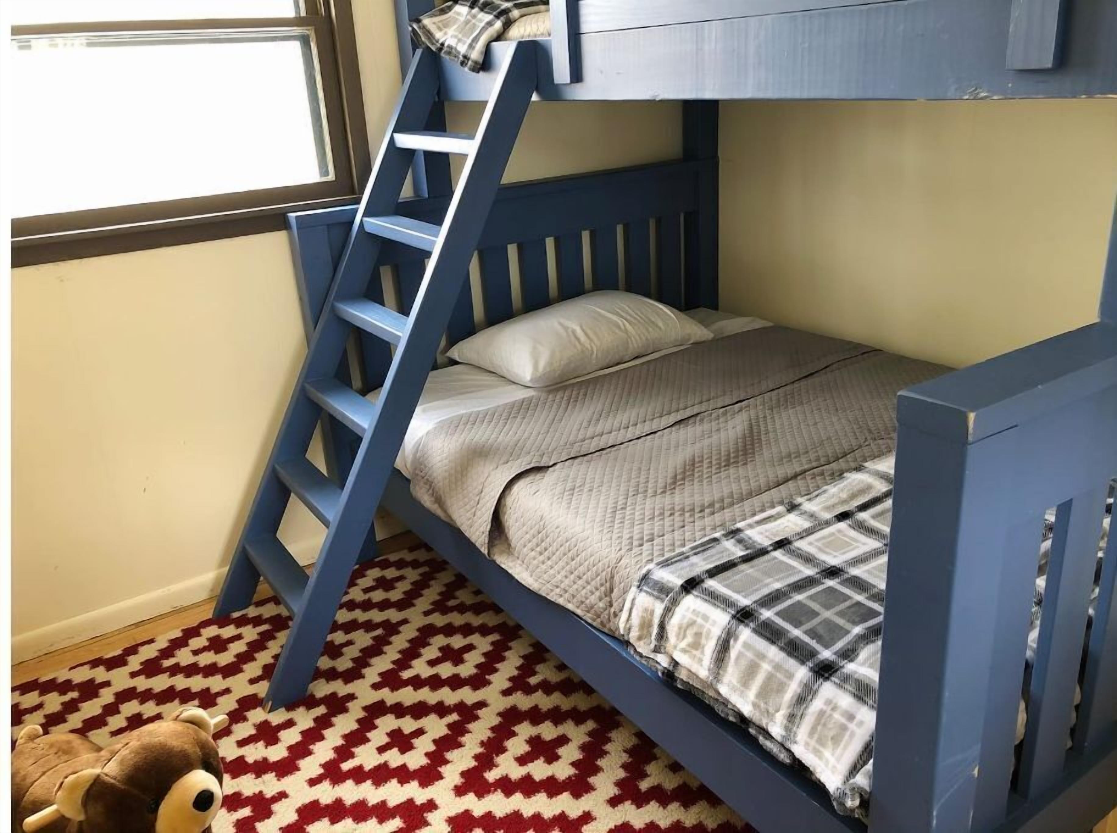 Un lit superposé avec une structure bleue, une échelle, un tapis à motifs et un jouet en peluche sur le sol.