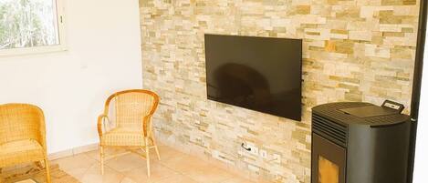A living room with a stone wall, a flat-screen TV, two wicker chairs, a wooden coffee table, and a wood-burning stove.