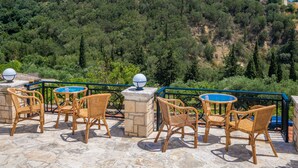 Outdoor wicker furniture with a stone floor, overlooking a hillside with dense trees and a clear blue sky. - Avrochares Villa (Ζάκυνθος)