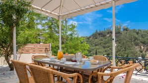 An outdoor dining area with wicker chairs and a table set for a meal, overlooking a mountainous landscape. - Avrochares Villa (Ζάκυνθος)