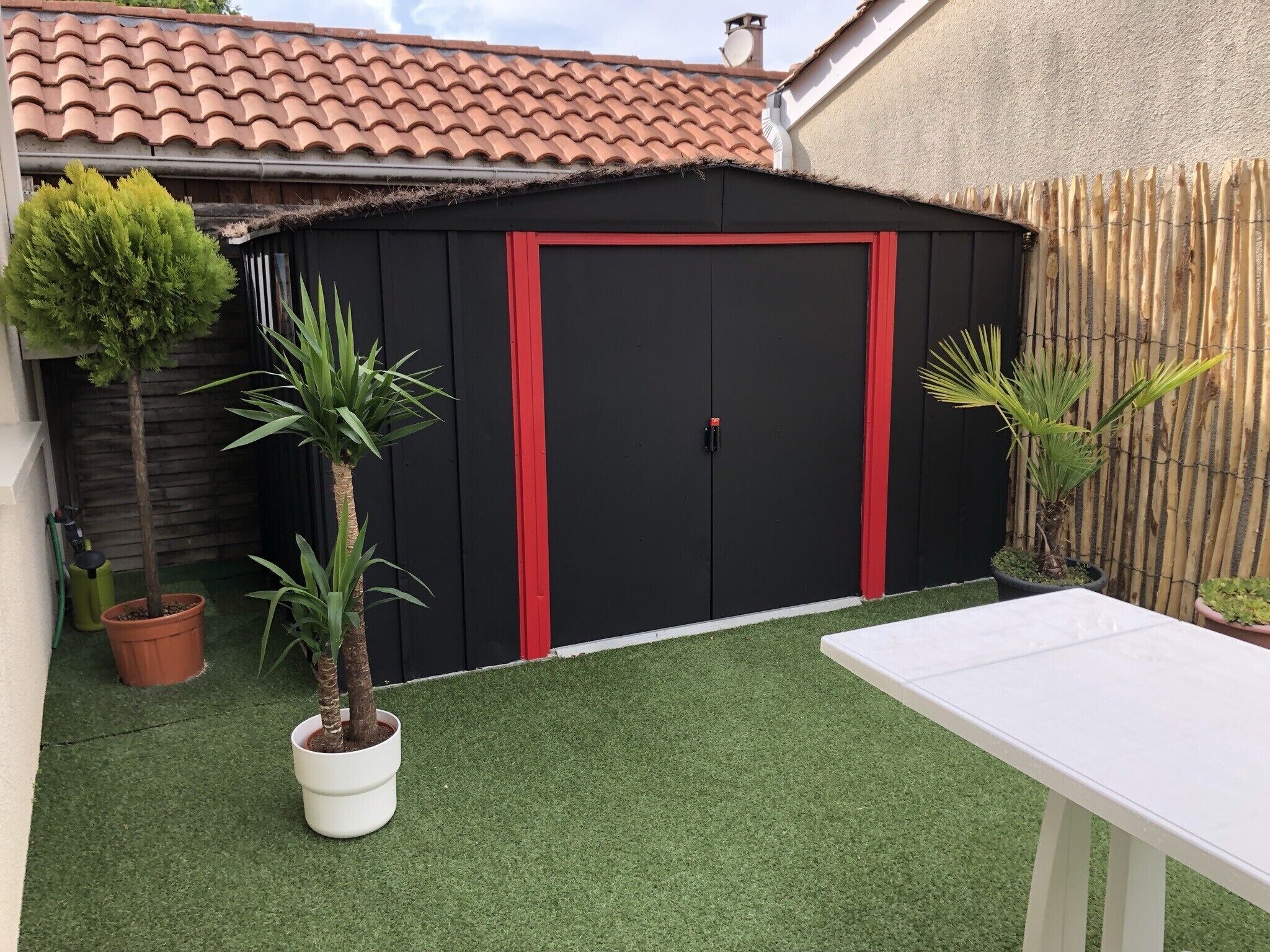 Un abri de jardin noir avec des bordures rouges, une table blanche et une pelouse artificielle verte.