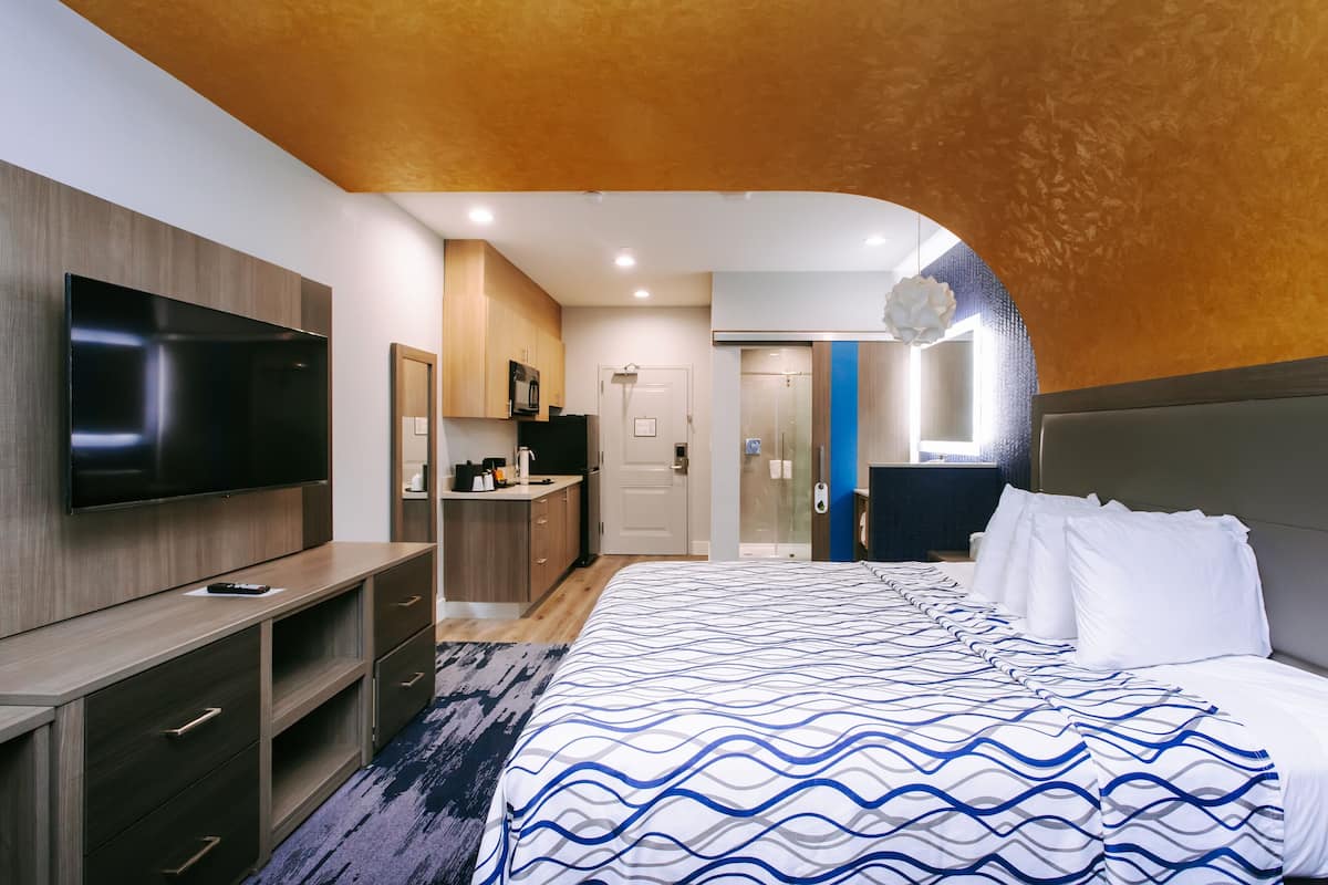 A modern hotel room with a large bed, a flat-screen TV, a kitchenette, and a bathroom visible through an arched doorway.