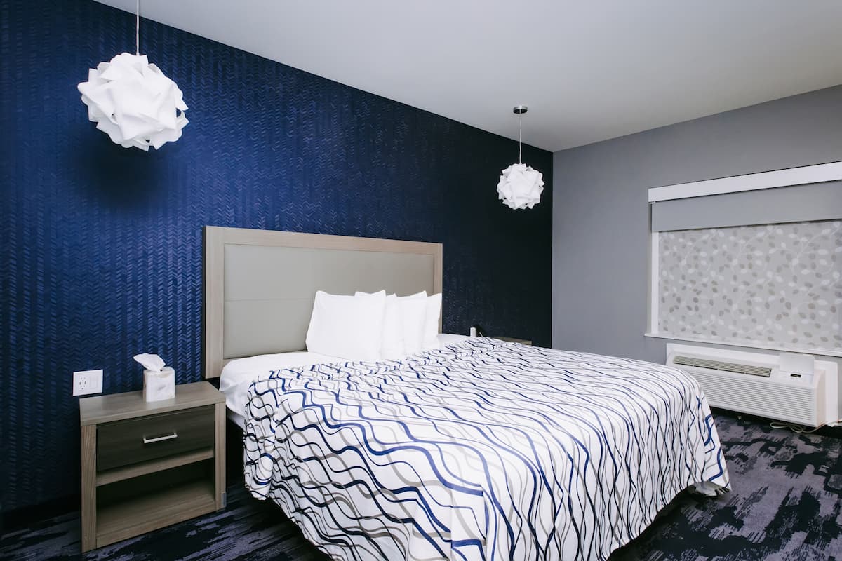 A hotel room with a bed, a nightstand, a wall-mounted heater, and a window with a patterned curtain.