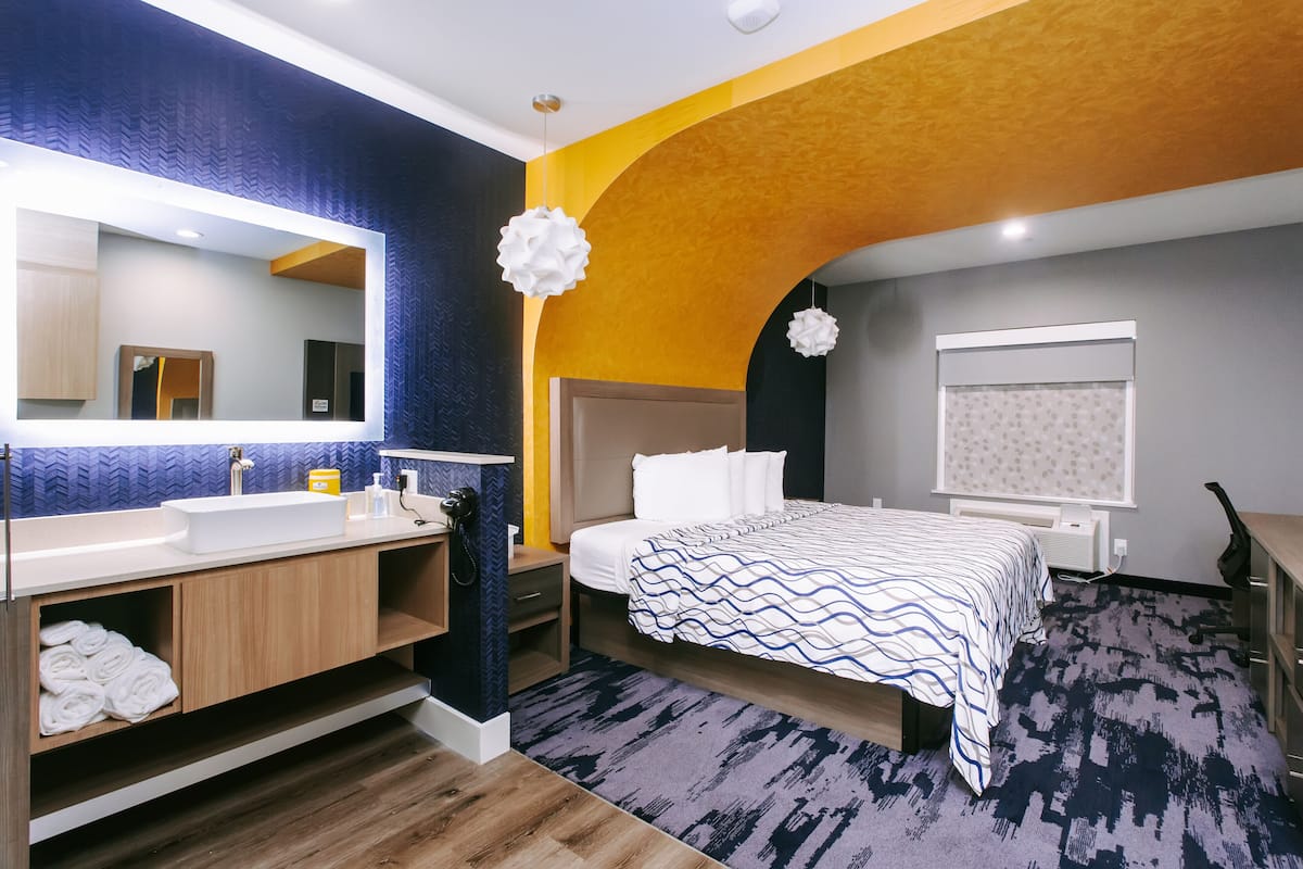 A modern hotel room with a large bed, a bathroom with a mirror and sink, and a unique ceiling design.