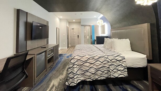 A modern hotel room with a large bed, a desk with a flat-screen TV, and a unique ceiling design. - Palace Inn Blue Houston East Beltway 8 (Houston)