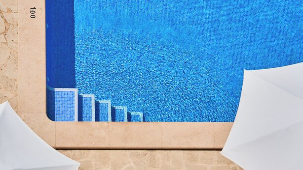 Una piscina con acqua cristallina e azzurra, circondata da piastrelle beige e un muro in pietra.