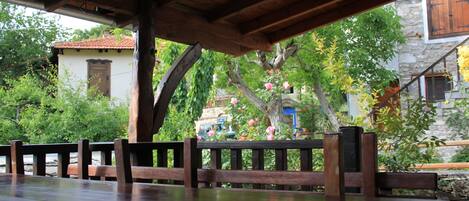 A wooden outdoor dining area with a table and chairs, surrounded by greenery and a stone building in the background.