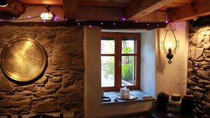 Een rustieke kamer met stenen muren, een houten plafond, een raam met uitzicht op groen, een stenen wandlamp en een ronde tafel met kaarsen en boeken.